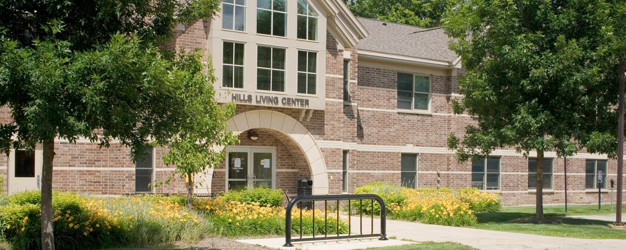 A brick building with large windows, labeled "Hills Living Center," surrounded by lush greenery and trees under a clear blue sky, exuding a peaceful vibe.